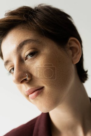 Nahaufnahme Porträt einer jungen sommersprossigen Frau mit Nasenpiercing, die isoliert auf grau in die Kamera blickt