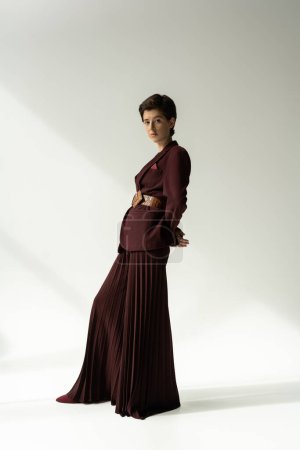 full length of young model in blazer and palazzo pants looking at camera on grey background with lighting