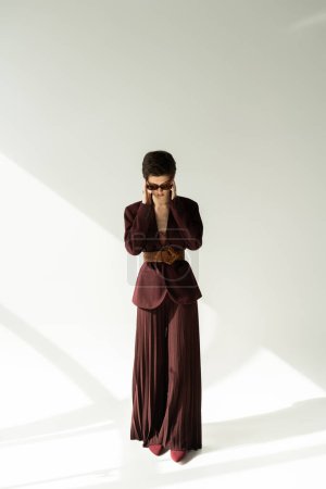 full length of brunette model in stylish jacket and palazzo pants adjusting sunglasses on grey background with lighting