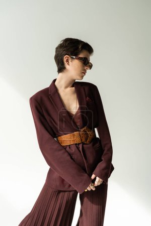 brunette woman in sunglasses and pantsuit with leather belt on grey background 