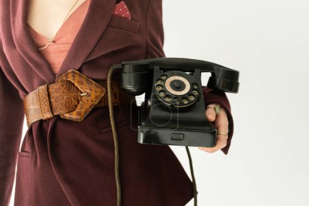 Ausgeschnittene Ansicht einer Frau in trendigem Blazer und Ledergürtel, die Vintage-Handy isoliert auf grau hält