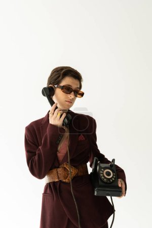 brunette model in sunglasses and fashionable blazer with leather belt calling on vintage phone isolated on grey