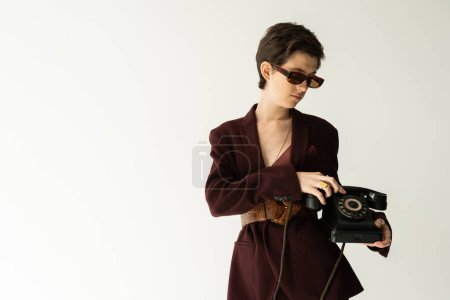 brunette woman in trendy jacket with leather belt standing with vintage phone on grey background