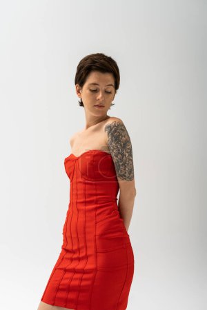 mujer tatuada de moda en vestido rojo sin tirantes posando con las manos detrás de la espalda aisladas en gris