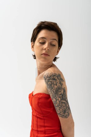 Foto de Mujer joven y encantadora con tatuaje posando en vestido de corsé rojo aislado en gris - Imagen libre de derechos