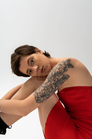 Foto de Mujer tatuada de ensueño en vestido de corsé rojo sentado y mirando a la cámara sobre fondo gris - Imagen libre de derechos