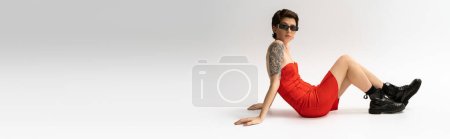 Photo for Full length of slim tattooed woman in black boots and red corset dress sitting on grey background, banner - Royalty Free Image