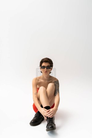 Foto de Longitud completa de mujer joven tatuada en botas negras ásperas y gafas de sol de moda sentadas sobre fondo gris - Imagen libre de derechos