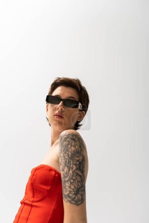 brünette tätowierte Frau mit Sonnenbrille und rotem Korsettkleid blickt in die Kamera auf grauem Hintergrund
