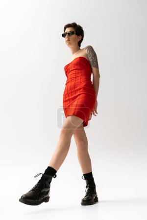 volle Länge der schlanken tätowierten Frau posiert in rotem Korsettkleid und schwarzen Stiefeln auf grauem Hintergrund