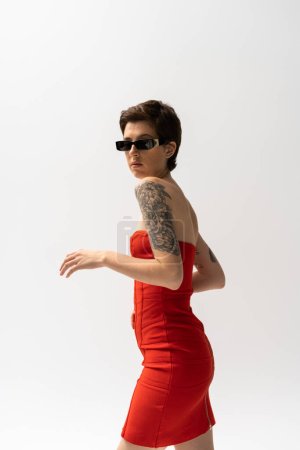 brünette tätowierte Frau mit Sonnenbrille und rotem Korsettkleid blickt vereinzelt in die Kamera auf grau