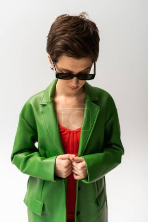 joven morena posando en gafas de sol de moda y chaqueta de cuero verde sobre fondo gris