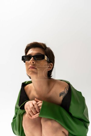 Téléchargez les photos : Femme tatouée dans une veste en cuir vert et des lunettes de soleil à la mode assis sur fond gris - en image libre de droit