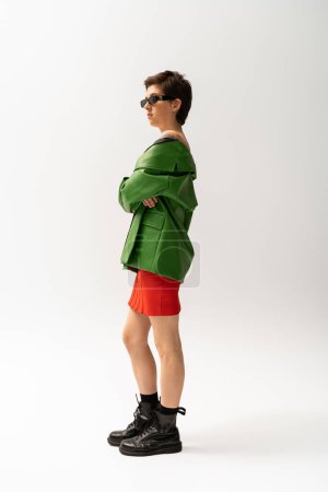Photo for Full length of stylish woman in green leather jacket and black boots standing with crossed arms on grey background - Royalty Free Image