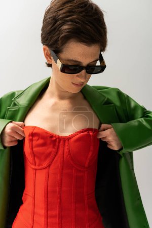 stylische Frau mit Sonnenbrille und grüner Lederjacke, dazu rotes trägerloses Kleid, isoliert auf grau