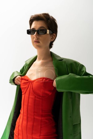 trendy woman in sunglasses and green leather jacket adjusting red corset dress on grey background
