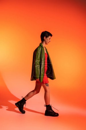 Foto de Vista lateral de mujer delgada en botas negras y chaqueta de cuero verde caminando sobre fondo naranja - Imagen libre de derechos