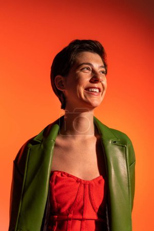 Foto de Alegre morena mujer en vestido de corsé rojo y chaqueta de cuero verde sonriendo y mirando hacia otro lado sobre fondo naranja - Imagen libre de derechos