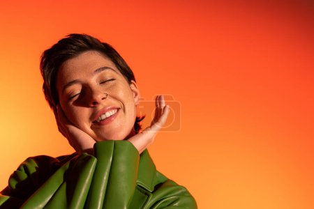 pleased brunette woman in green leather jacket holding hands near face and smiling with closed eyes on orange background
