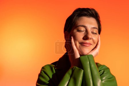 happy brunette woman with closed eyes touching face and smiling on orange background