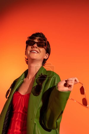 excited and fashionable woman in green leather jacket posing with different sunglasses on orange background