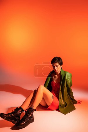 volle Länge der modischen brünetten Frau in grüner Jacke und schwarzen Lederstiefeln sitzt auf orangefarbenem Hintergrund