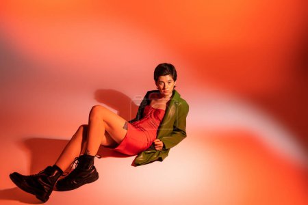 Photo for Full length of tattooed woman in red corset dress and green jacket with black boots lying on colorful background - Royalty Free Image