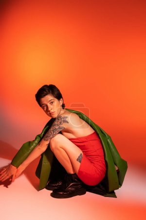 longitud completa de mujer tatuada en vestido de corsé rojo y botas negras sentadas en las horquillas y mirando a la cámara sobre fondo naranja y rosa