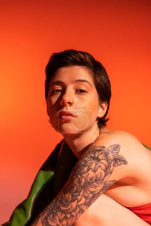 Téléchargez les photos : Portrait de femme coûteuse avec tatouage et cheveux bruns courts regardant la caméra sur fond orange - en image libre de droit