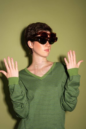 Photo for Brunette woman in striped jumper wearing several trendy sunglasses on green background - Royalty Free Image