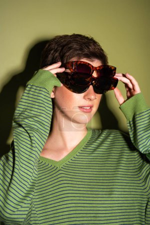 young woman in trendy striped pullover wearing several sunglasses on green background