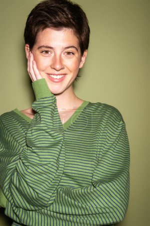 portrait of joyful brunette woman in striped pullover touching face and looking at camera on green background