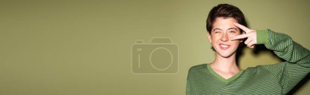 femme brune souriante clignant des yeux à la caméra et montrant signe de paix près du visage sur fond vert, bannière