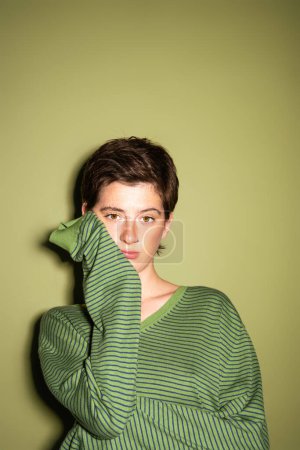 thoughtful brunette woman in trendy pullover looking at camera on green background