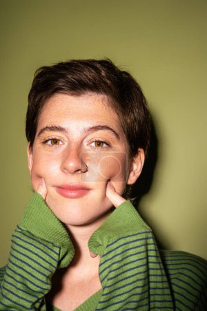 portrait of freckled brunette woman in spring jumper touching cheeks on green background