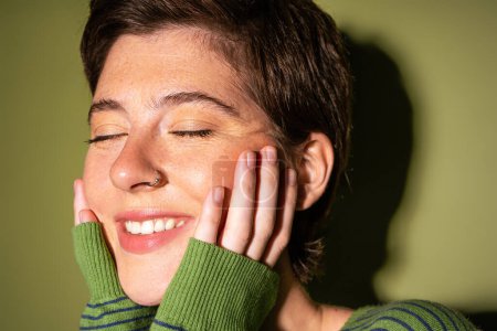 Porträt einer überglücklichen Frau mit Sommersprossen und Nasenpiercing, die das Gesicht berührt und mit geschlossenen Augen auf grünem Hintergrund lächelt