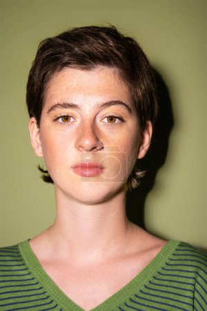 Porträt einer brünetten Frau mit Sommersprossen und Nasenpiercing, die in die Kamera auf grünem Hintergrund blickt