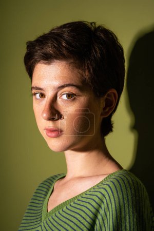 portrait of young freckled woman with short brunette hair and nose piercing looking at camera on green background with shadow