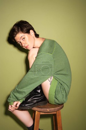 Nachdenkliche Frau in gestreiftem Pullover und schwarzen Lederstiefeln sitzt auf einem Holzhocker auf grünem Hintergrund