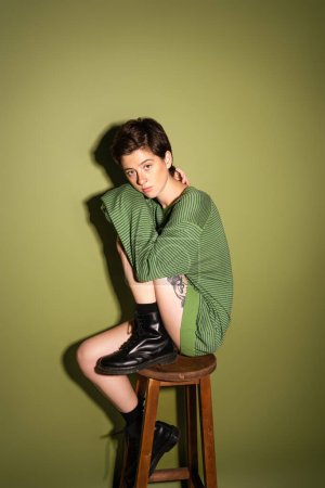 volle Länge der trendigen Frau in gestreiftem Pullover und schwarzen Lederstiefeln sitzt auf einem Holzhocker auf grünem Hintergrund mit Schatten