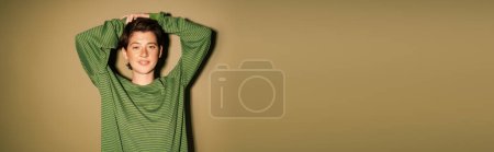 fröhliche Frau im gestreiften Pullover posiert mit den Händen über dem Kopf und lächelt in die Kamera auf grünem Hintergrund, Banner
