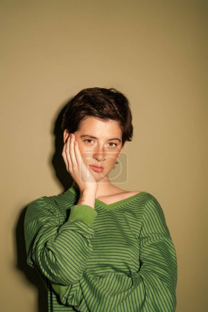 displeased brunette woman in striped jumper looking at camera while holding hand near face on green background