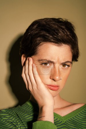 portrait of upset freckled woman with short brunette hair touching face and looking at camera on green background