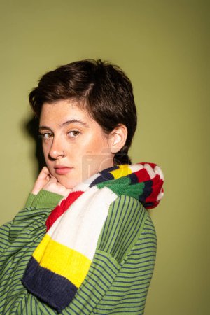 portrait of dreamy brunette woman in striped multicolored scarf looking at camera on green background