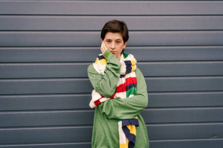 skeptical woman in green pullover and striped multicolored scarf posing with hand near face at grey wall on street