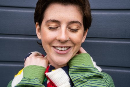 portrait of stylish freckled woman smiling with closed eyes near grey wall on street
