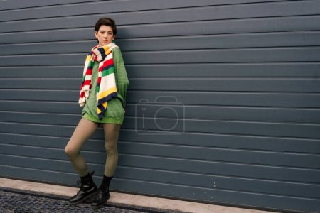 Photo for Full length of woman in striped scarf and black leather boots posing near grey wall on street - Royalty Free Image