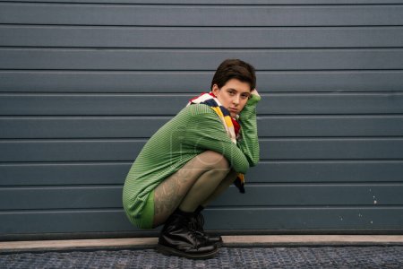 full length of trendy woman in green pullover and striped scarf sitting on haunches near grey wall on street