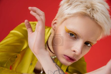blonde albino woman with bright eyeliner and tattoo looking at camera while posing on carmine pink 