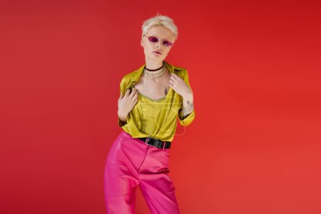 tattooed albino model in stylish outfit and trendy sunglasses posing on carmine pink background 